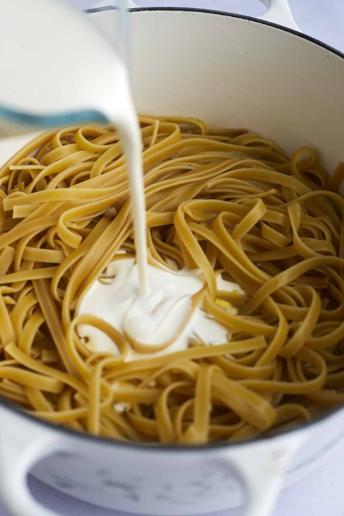 alfredo sauce being poured over cooked fettuccine noodles in a pot 