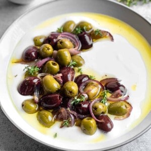 a white bowl full of whipped feta topped with kalamata and green roasted olives, red onions, and a drizzle of olive oil