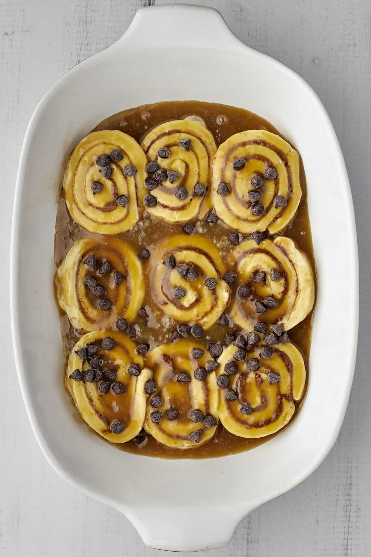 unbaked cinnamon rolls layered between caramel sauce topped with chocolate chips in a white baking dish