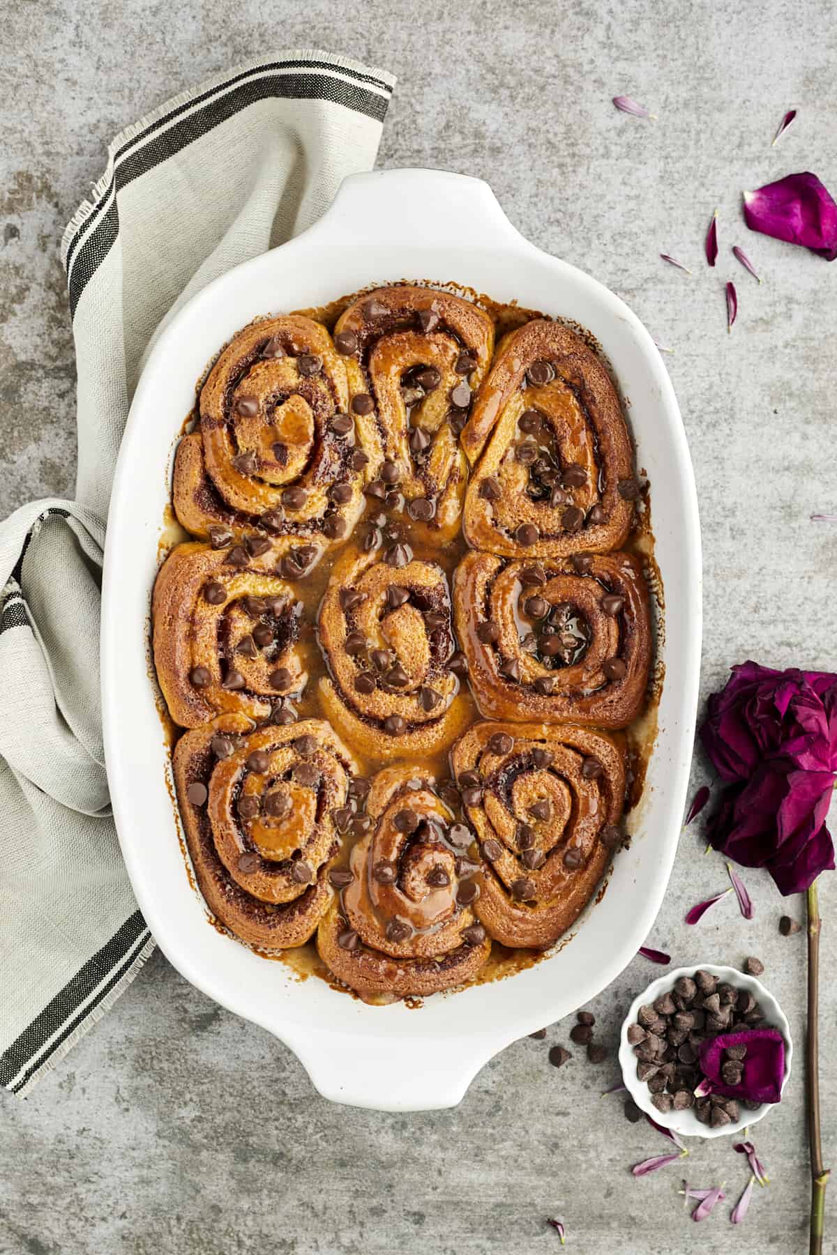 overhead image of easy cinnamon rolls topped with caramel and chocolate chips in a white baking dish with a towel and a small bowl of chocolate chips off to the side