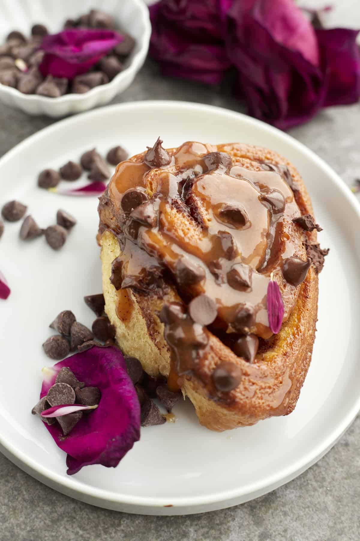 a caramel chocolate chip cinnamon roll on a white plate with scattered chocolate chips and a pink rose