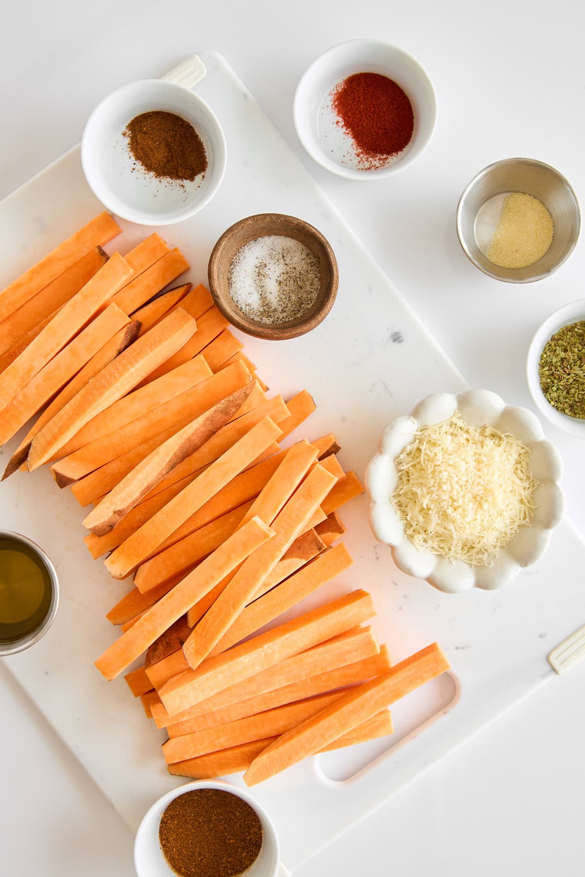 Raw sweet potato fries on a cutting board next to bowls of spices.