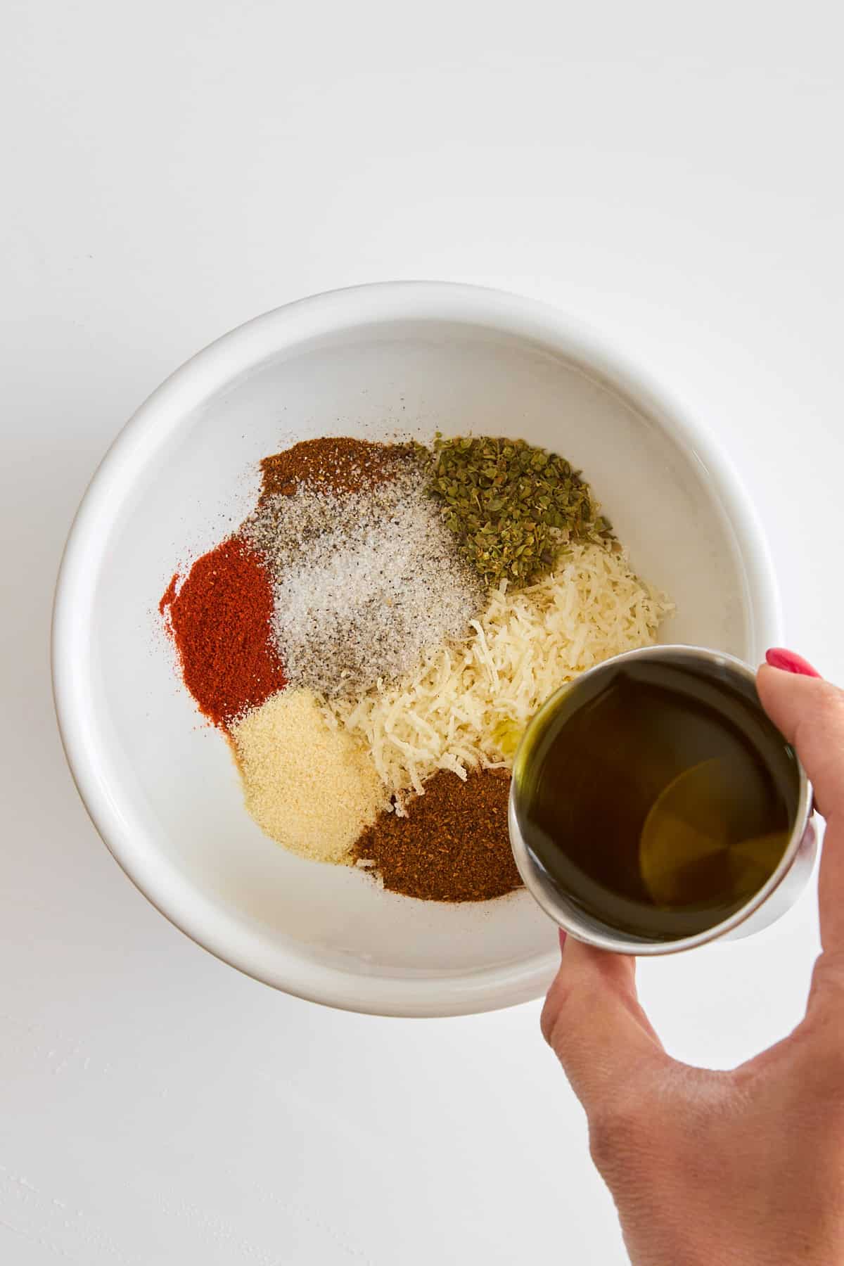 A bowl of seasonings and cheese and a hand holding olive oil on top.