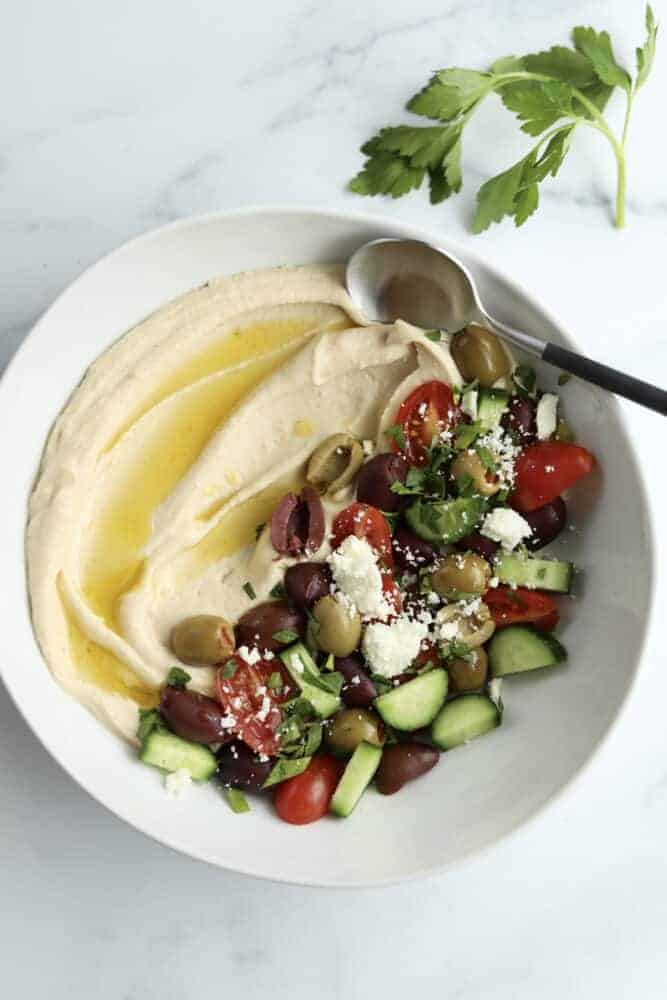a bowl of hummus with roasted tomatoes and olives with a spoon sticking out