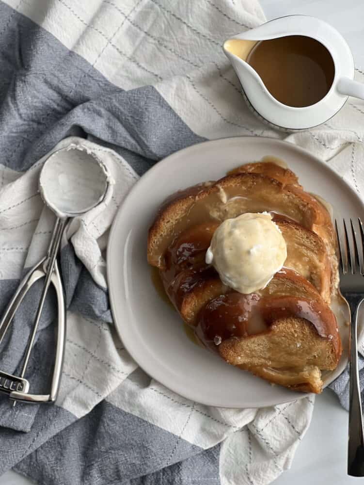 a serving of salted caramel bread pudding on a plate topped with vanilla ice cream with a fork on the plate and an ice cream scoop to the left