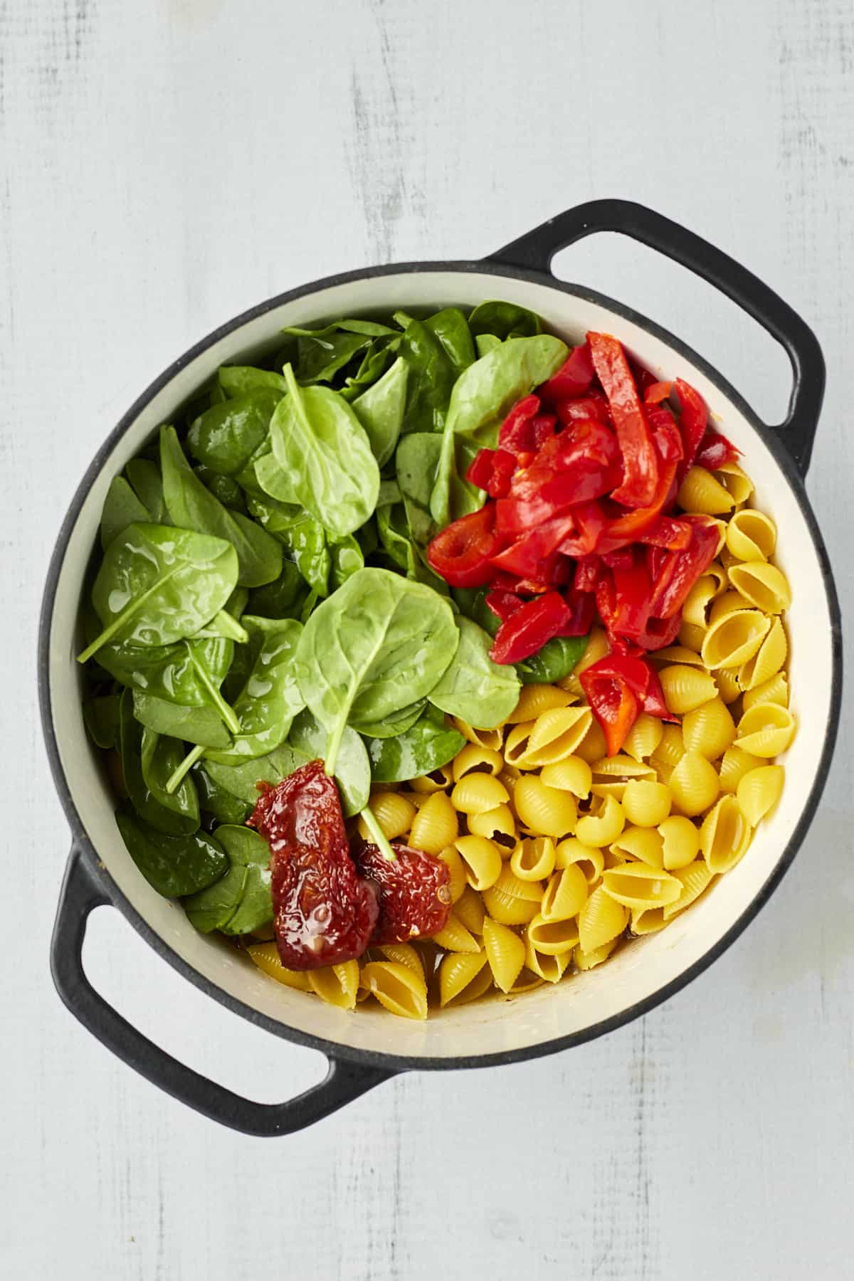 Cooked pasta shells, red bell peppers, spinach, and roasted tomatoes in a large pot.