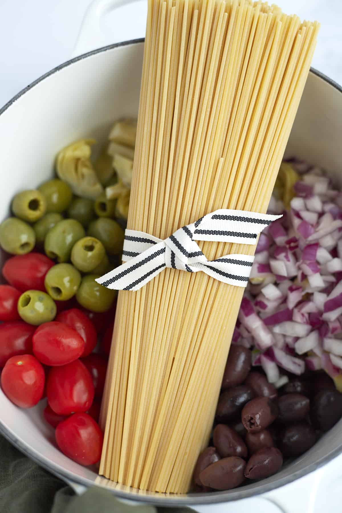 green and black olives, cherry tomatoes, chopped red onion, and a bundle of spaghetti noodles in a pot 