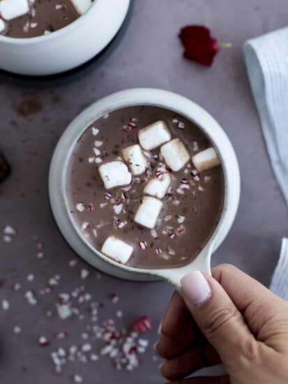 a hand holding a mug of hot chocolate topped with marshmallows and peppermint pieces