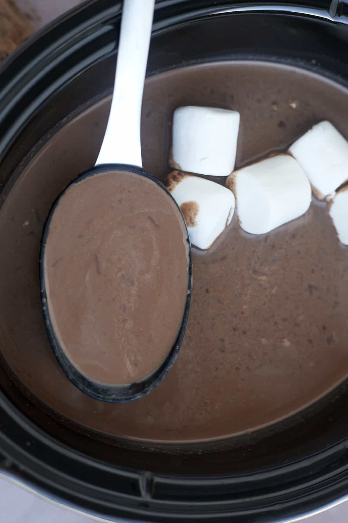 a spoon scooping up hot chocolate from a slow cooker with marshmallows floating on top
