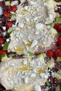close up image of three raw chicken breasts topped with hummus, cheese, and oil surrounded by vegetables
