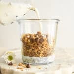 A hand pours milk from a glass pitcher into a clear jar filled with granola, chocolate chips, and chia seeds, sitting on a marble board with a small white flower nearby.