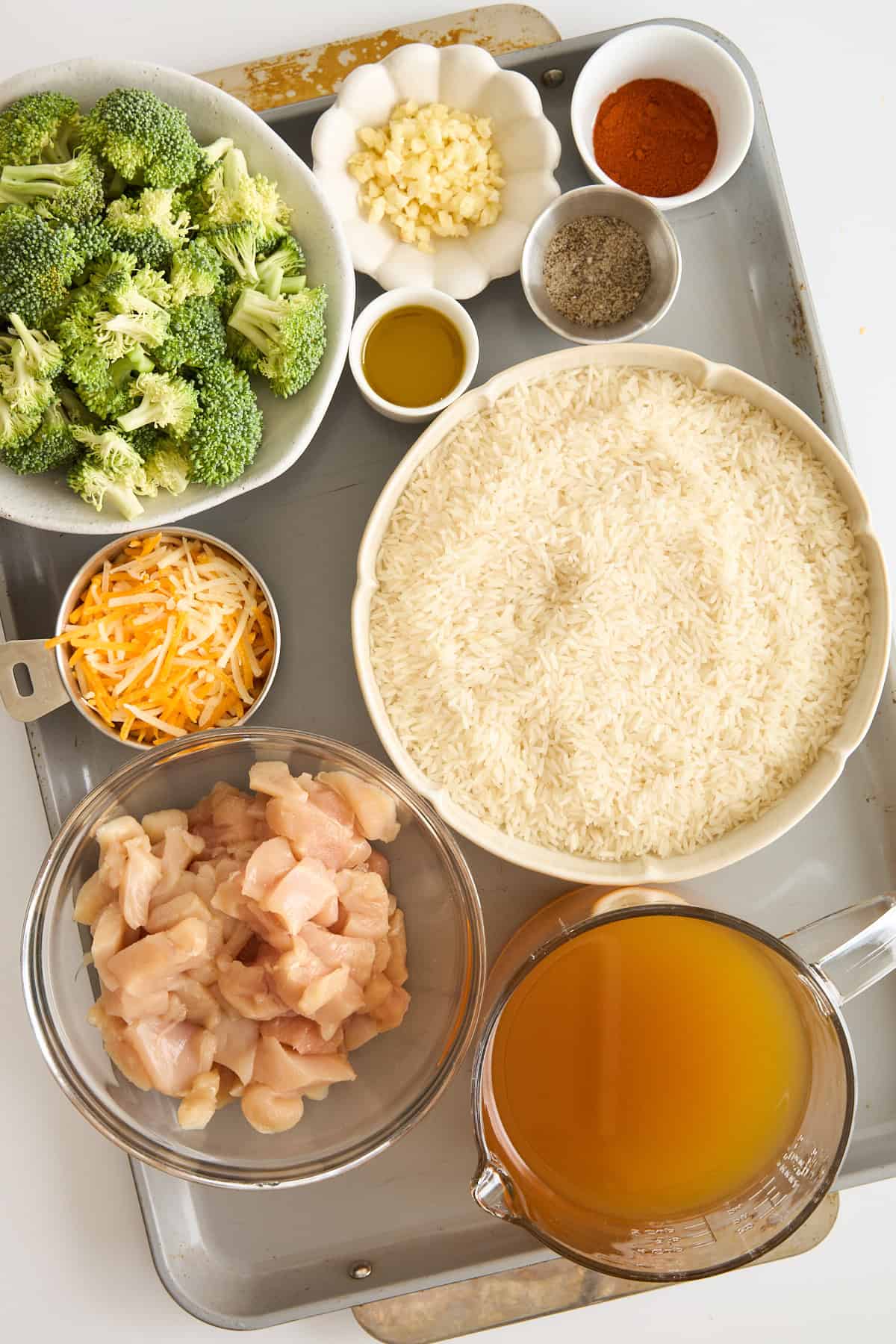 An overhead view of a tray holding bowls of raw broccoli, chopped garlic, shredded cheese, uncooked rice, diced raw chicken, chicken broth, olive oil, and small dishes of spices.