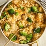 A skillet filled with rice, broccoli florets, and pieces of chicken, with a gold serving spoon resting inside. A striped cloth and stacked plates with forks are nearby on a light surface.