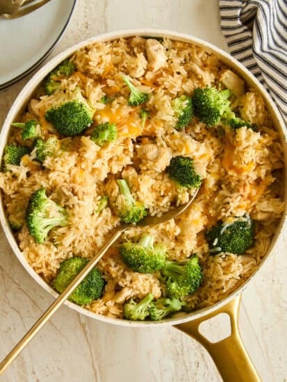 A skillet filled with rice, broccoli florets, and pieces of chicken, with a gold serving spoon resting inside. A striped cloth and stacked plates with forks are nearby on a light surface.