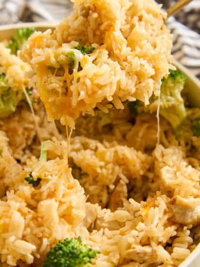 A close-up of cheesy rice with broccoli and chunks of chicken being scooped out of a dish, with melted cheese stretching from the spoon.