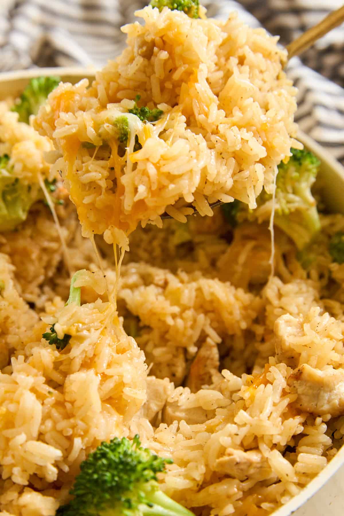 A close-up of cheesy rice with broccoli and chunks of chicken being scooped out of a dish, with melted cheese stretching from the spoon.
