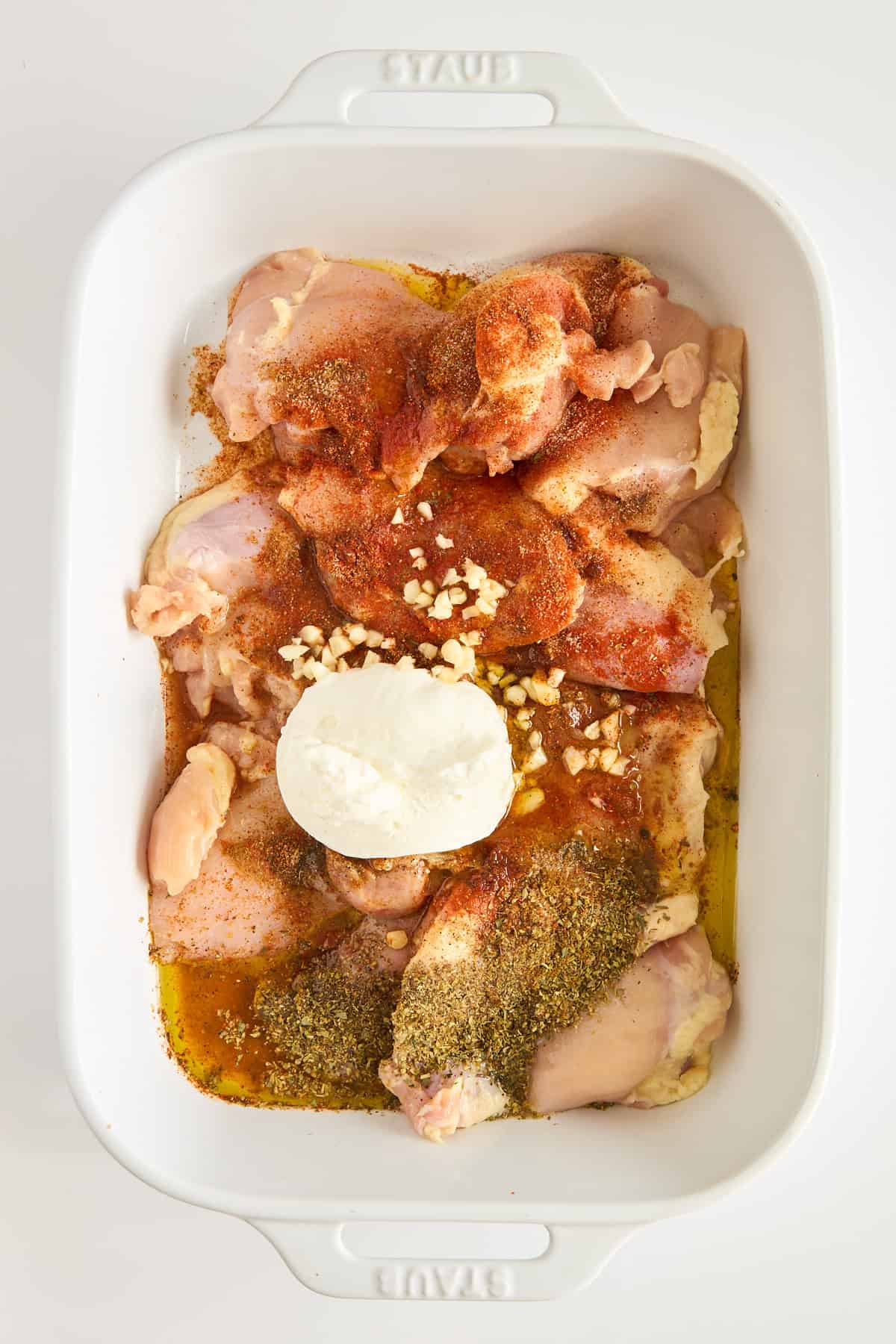 Raw chicken thighs in a white baking dish, topped with minced garlic, herbs, paprika, ground spices, and a dollop of sour cream or yogurt, ready to be mixed or marinated.