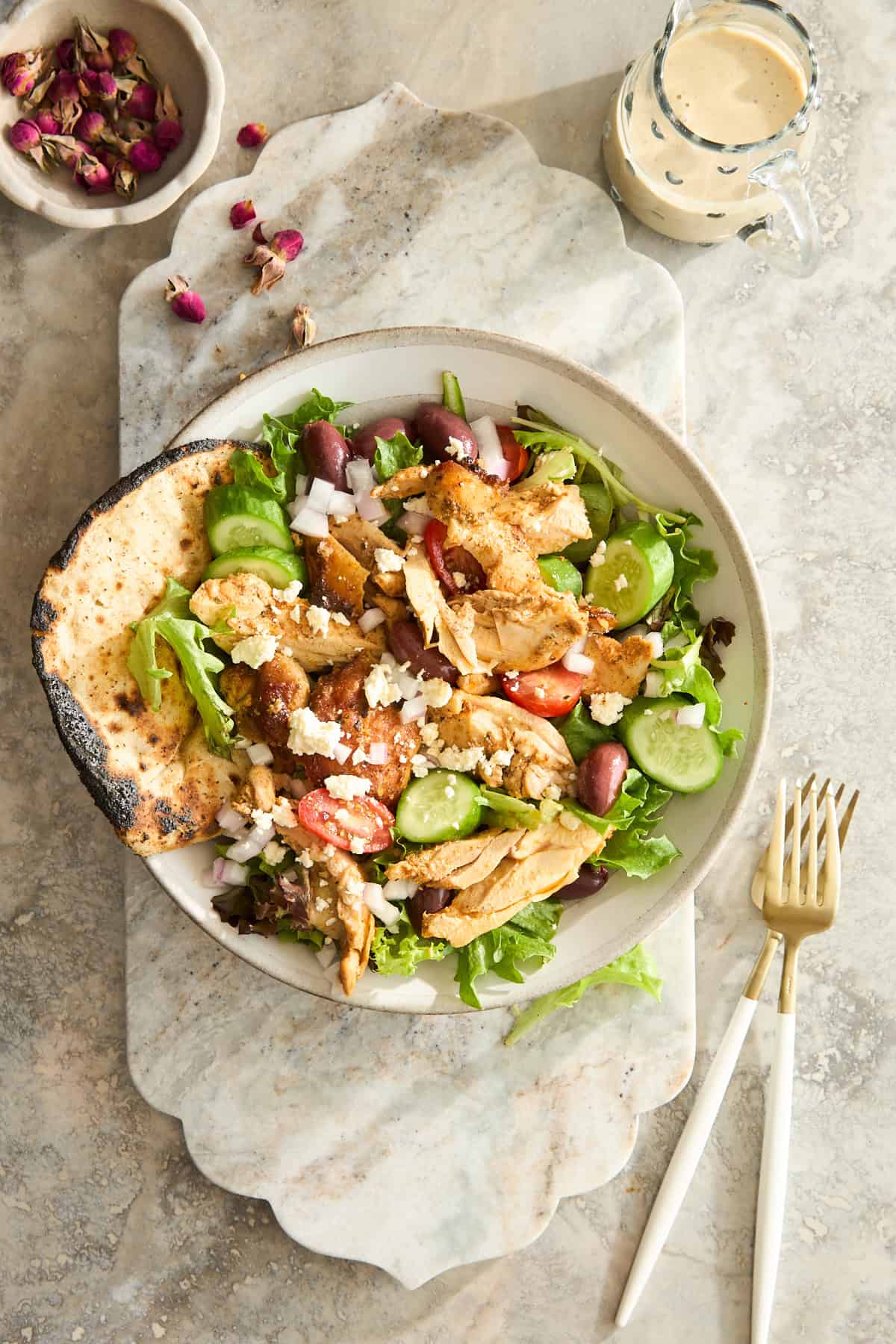 A bowl of salad with grilled chicken, cucumber, cherry tomatoes, olives, onions, feta cheese, and mixed greens, served with a piece of flatbread. A small jar of dressing and cutlery are beside the bowl on a marble surface.
