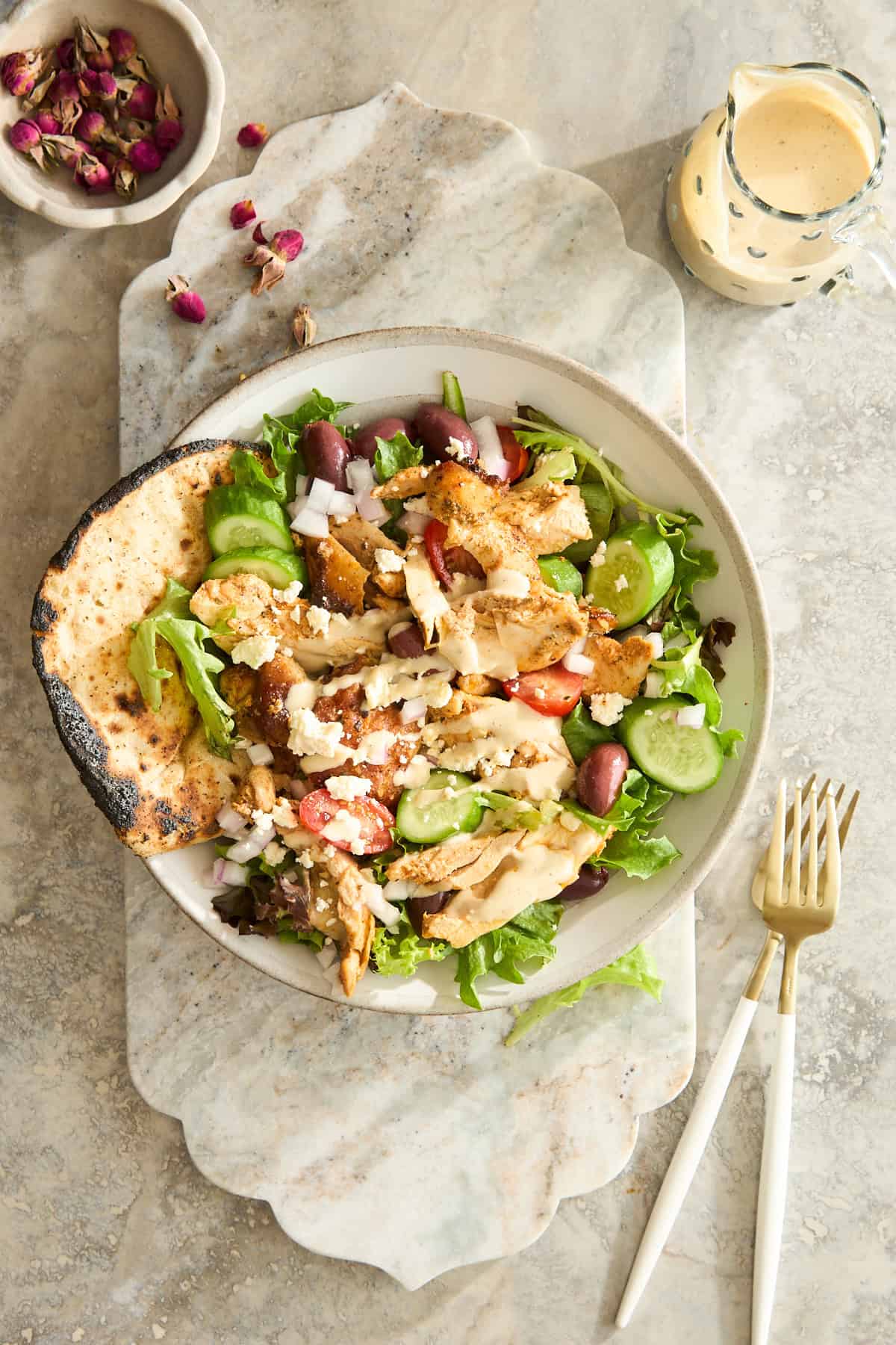 A bowl of Greek salad with grilled chicken, pita bread, cucumbers, tomatoes, feta cheese, and olives on a marble surface, with a fork, knife, a small pitcher of dressing, and a bowl of dried rose petals nearby.