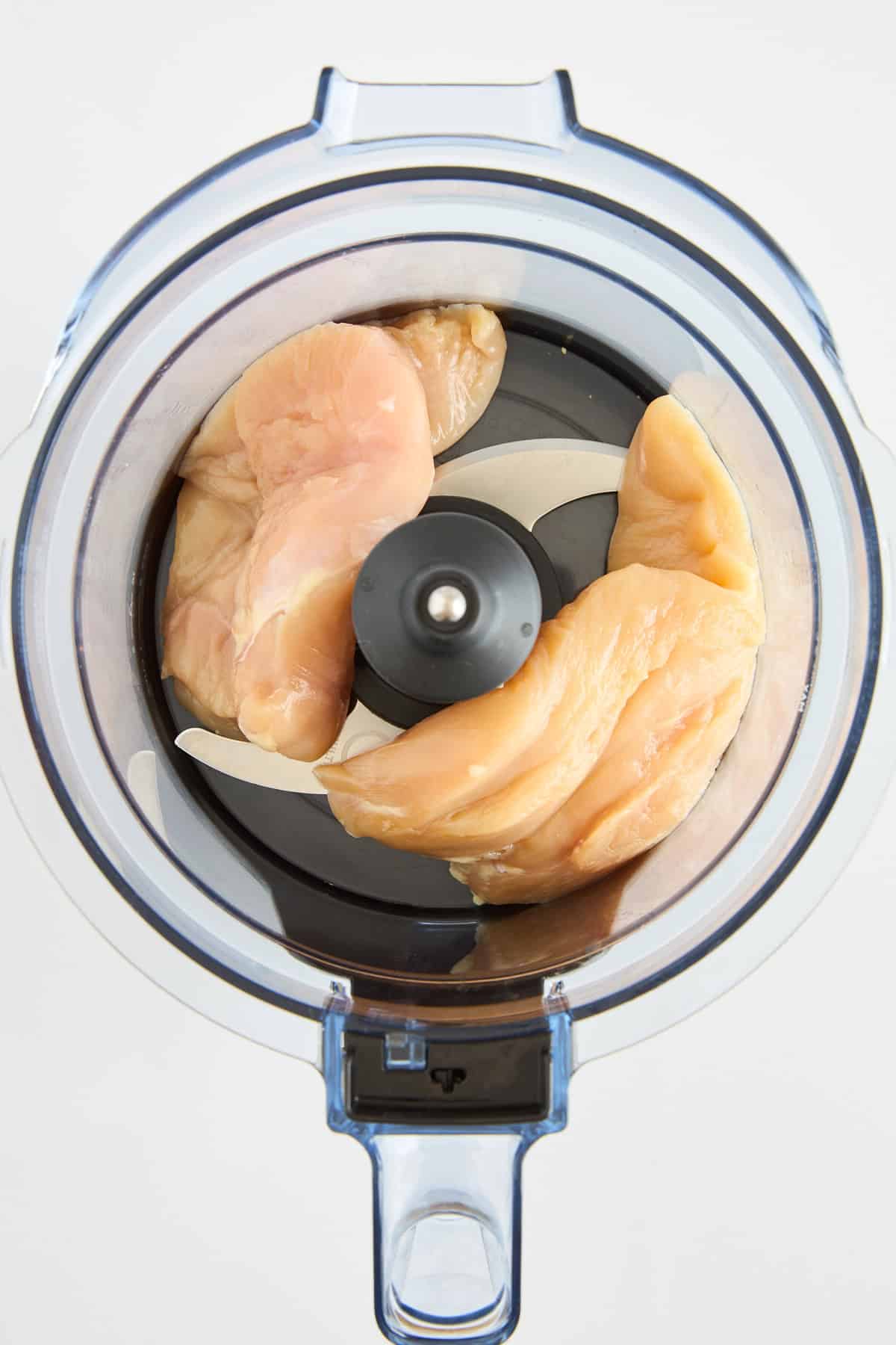 Raw chicken breast pieces placed inside a food processor, viewed from above, ready to be processed. The food processor has a clear plastic bowl and a sharp metal blade in the center.