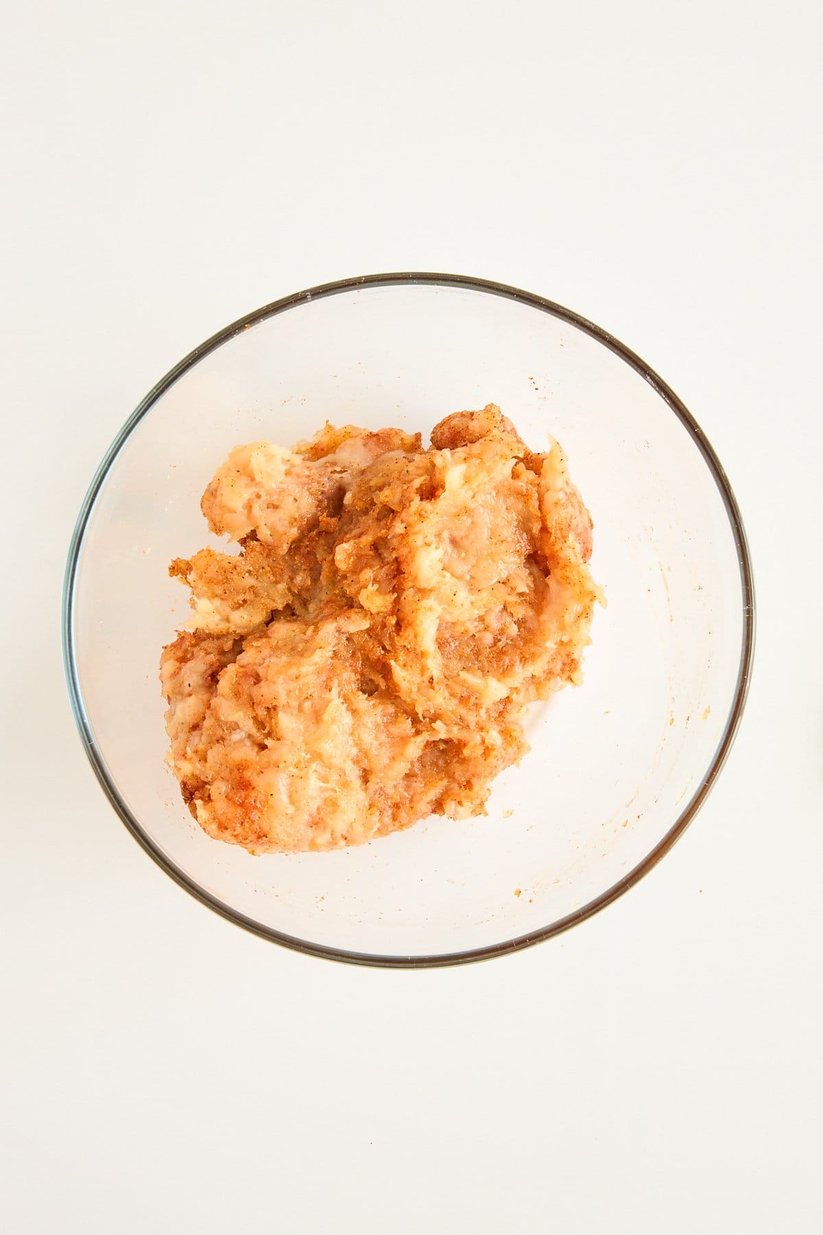 A clear glass bowl containing a mixture of ground meat and seasonings on a white surface.