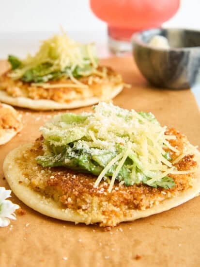 A close-up of a flatbread topped with green lettuce, shredded cheese, and a sprinkle of grated cheese, with another similar flatbread and a pink drink blurred in the background.