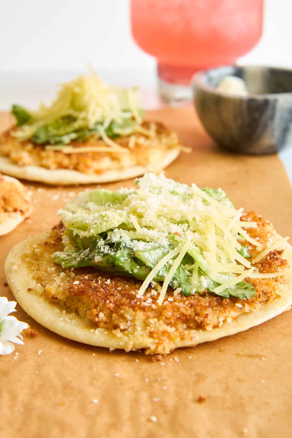 A flatbread topped with green lettuce, shredded cheese, and a sprinkle of grated cheese, with another similar flatbread and a pink drink blurred in the background.