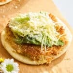 A crispy round flatbread topped with fresh green lettuce, grated cheese, and a sprinkle of parmesan, served on brown parchment paper with a white daisy flower beside it.