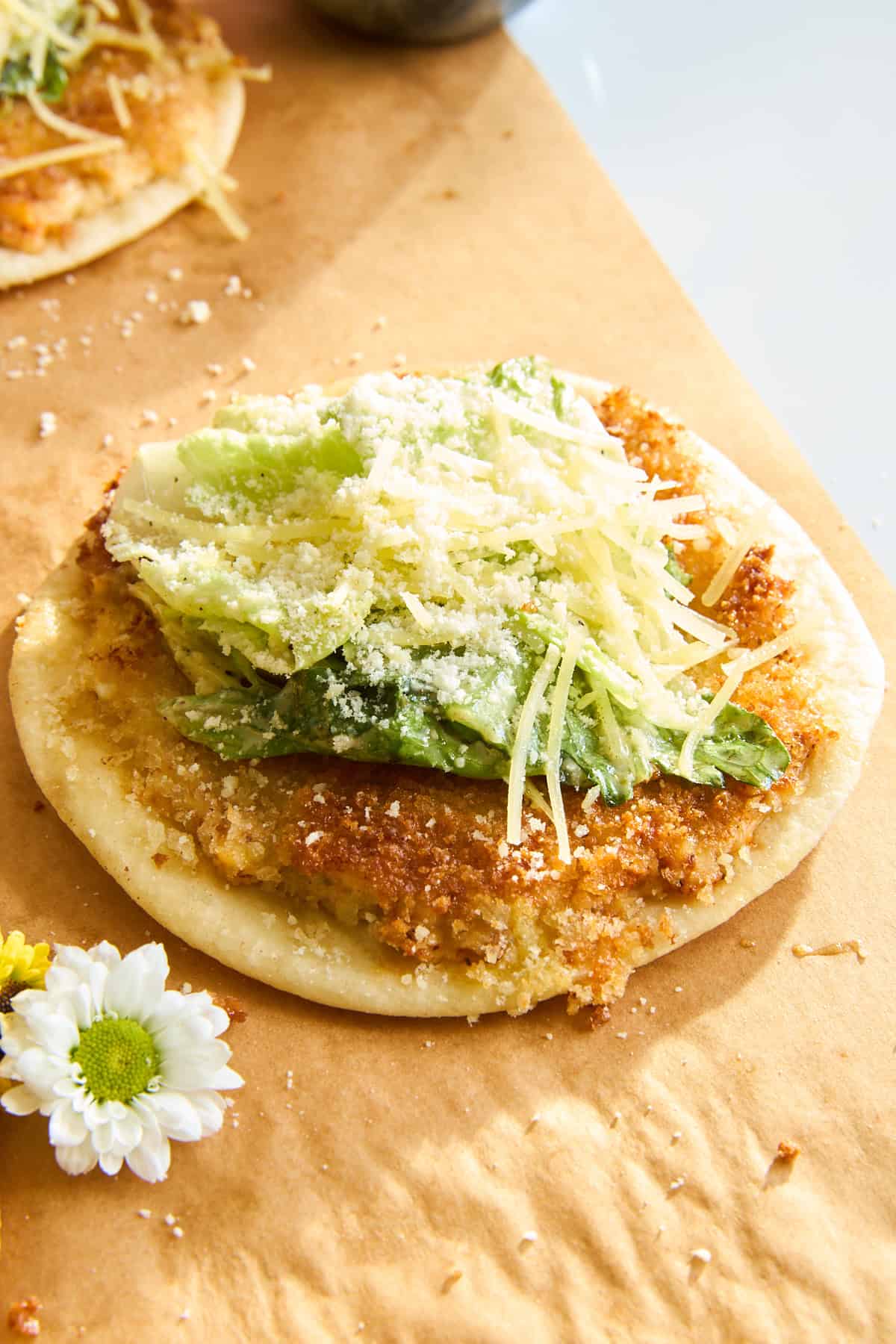 A crispy round flatbread topped with fresh green lettuce, grated cheese, and a sprinkle of parmesan, served on brown parchment paper with a white daisy flower beside it.