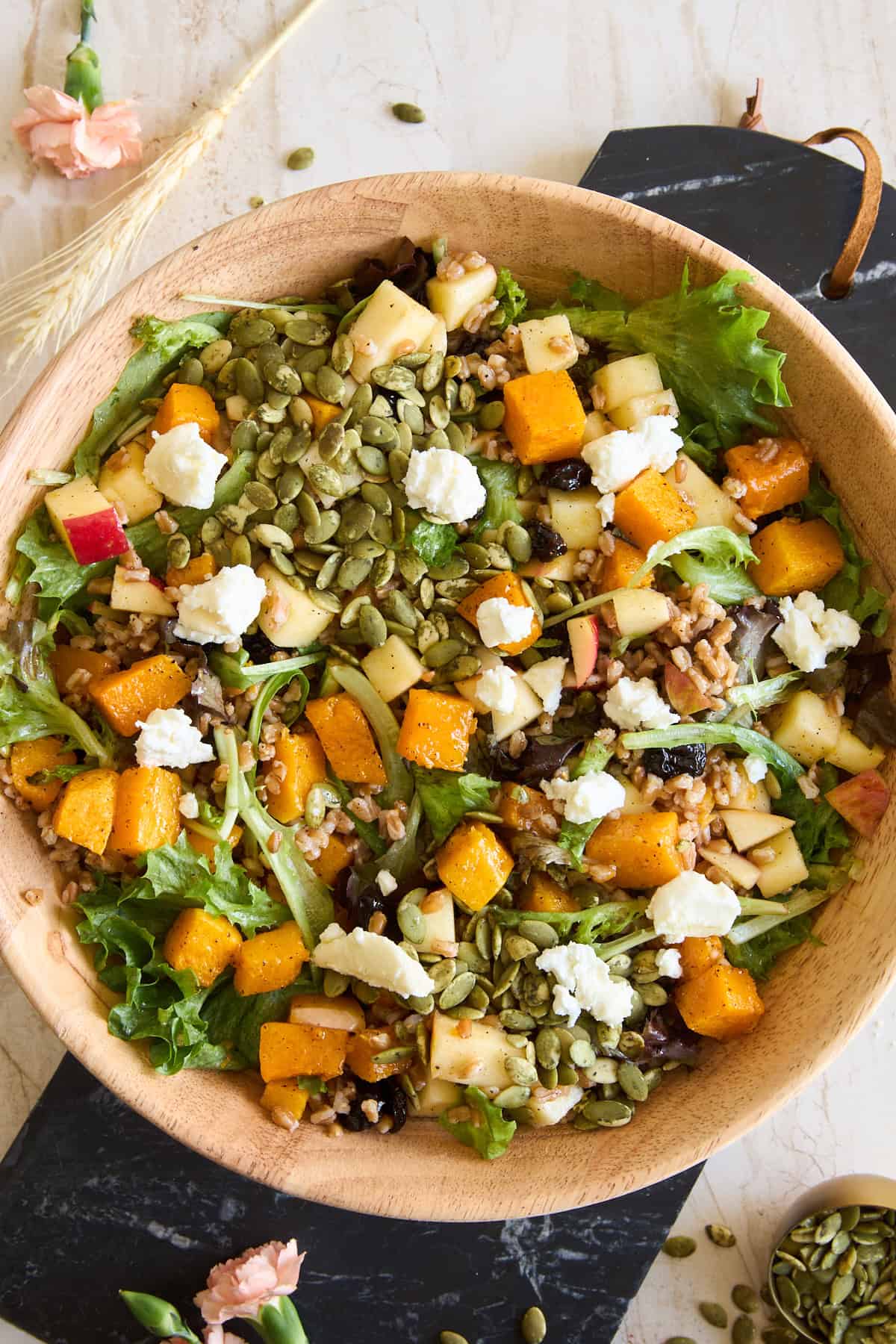A wooden bowl filled with mixed greens, roasted butternut squash, diced apples, goat cheese crumbles, pumpkin seeds, and grains on a light table with a black slate and flowers nearby.