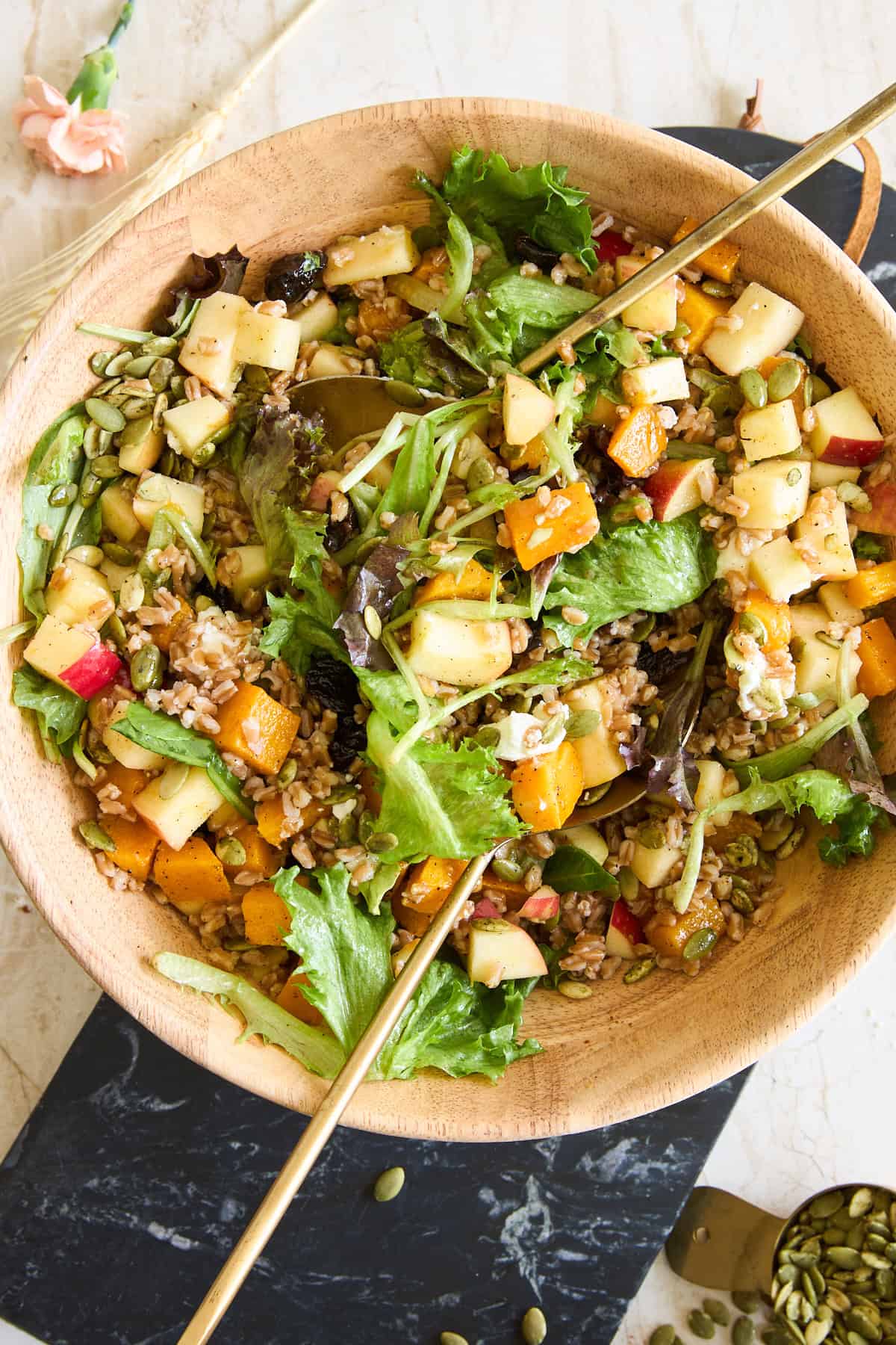 A wooden bowl filled with a mixed salad of leafy greens, chopped apples, roasted butternut squash, seeds, and grains, with two gold utensils. The bowl sits on a dark board with seeds scattered nearby.