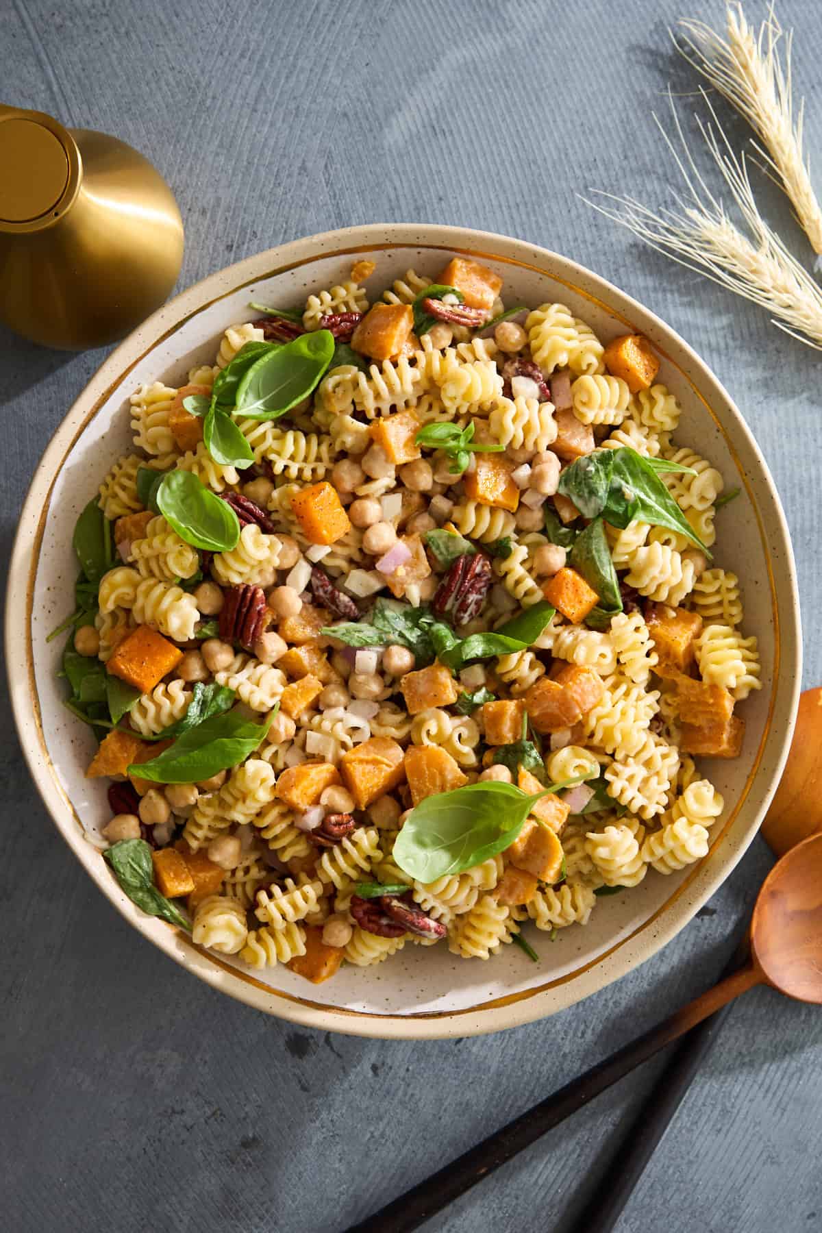 A bowl of pasta salad with chickpeas, roasted sweet potatoes, pecans, and fresh spinach leaves, garnished with basil, set on a grey table with a gold pepper grinder and wooden utensils nearby.