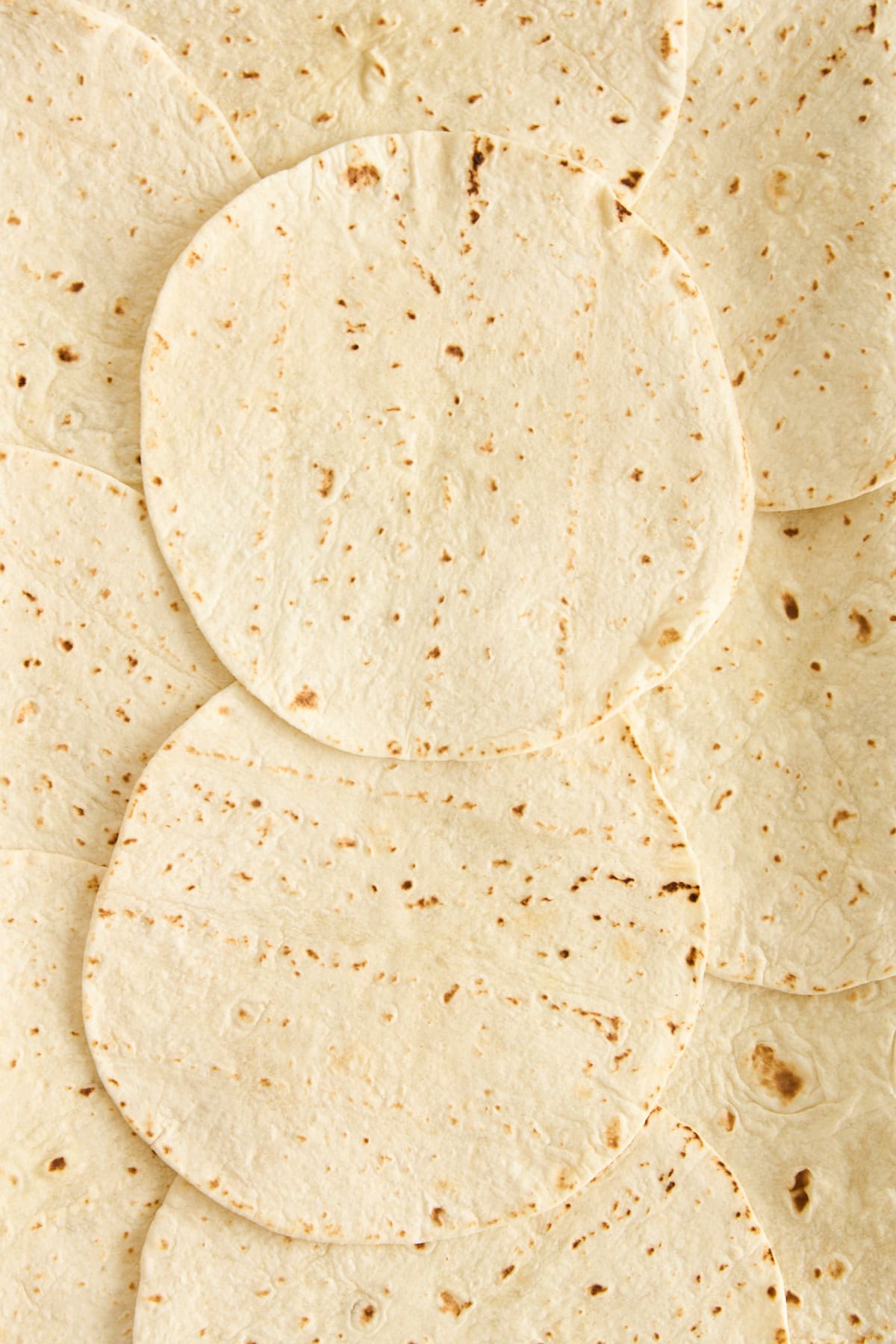 Multiple flour tortillas are laid out overlapping each other, covering the entire surface. The tortillas have light brown spots and a soft, slightly textured appearance.