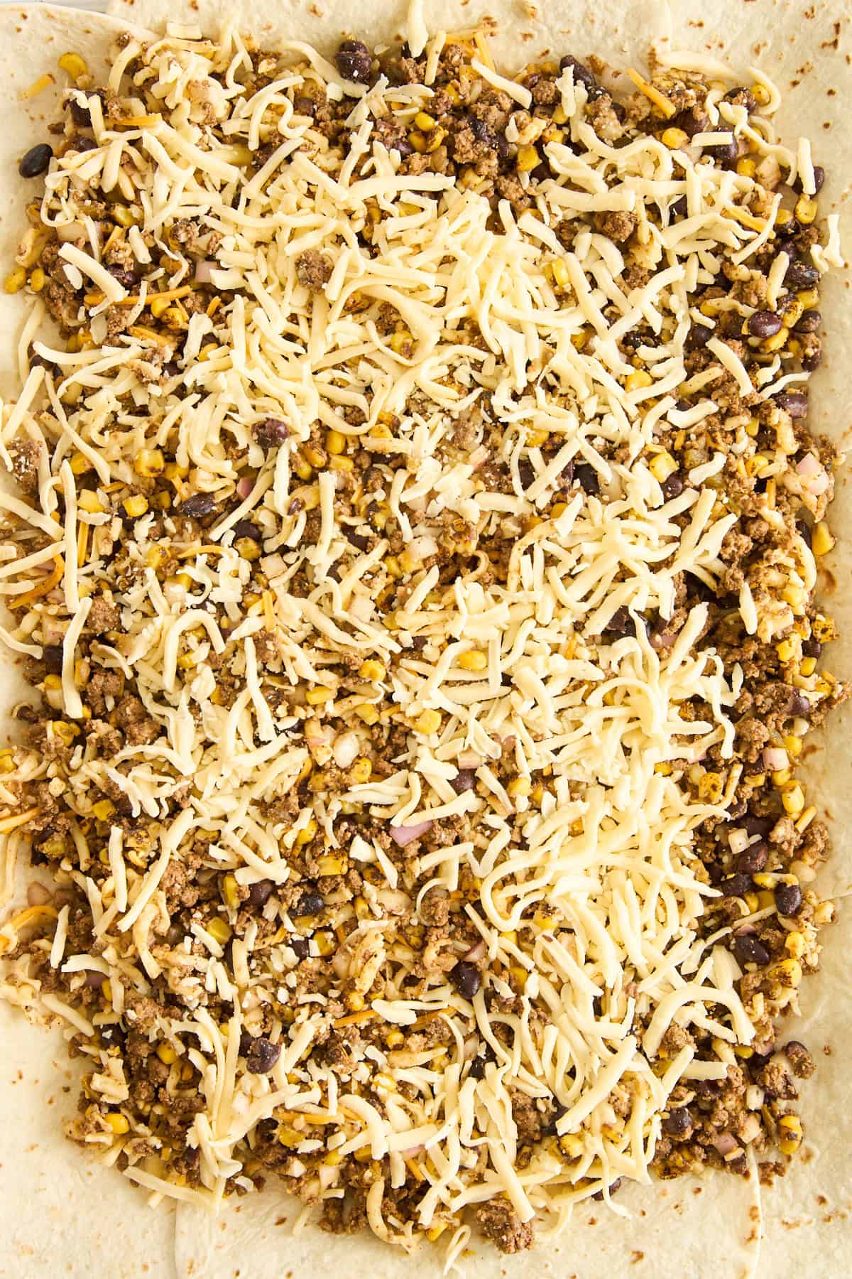 A close-up view of a flour tortilla topped with shredded cheese, cooked ground meat, black beans, corn, and rice, ready to be rolled or folded for a burrito or wrap.