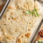 A baking sheet covered with overlapping toasted flour tortillas, garnished with fresh basil. Small bowls of sour cream and salsa sit nearby on a beige surface.