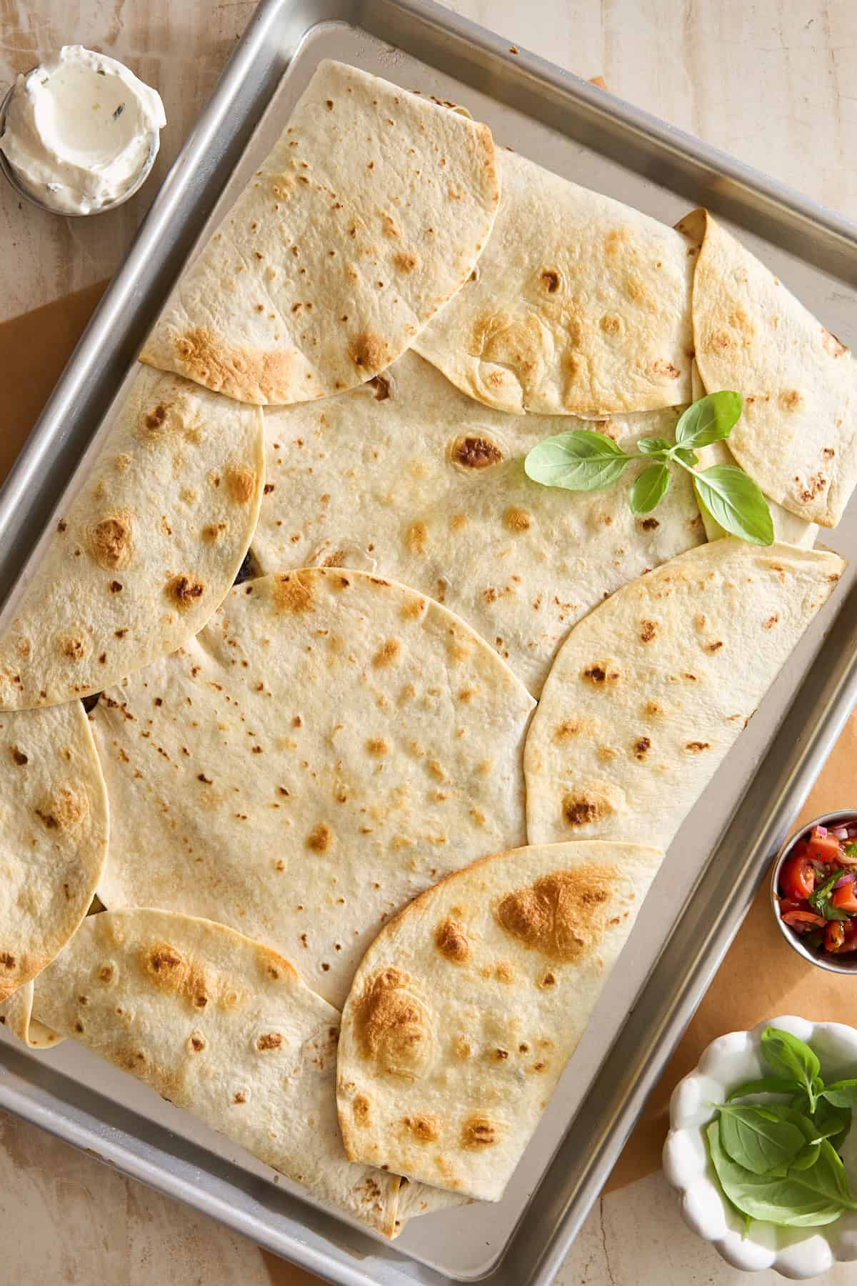 A baking sheet covered with overlapping toasted flour tortillas, garnished with fresh basil. Small bowls of sour cream and salsa sit nearby on a beige surface.