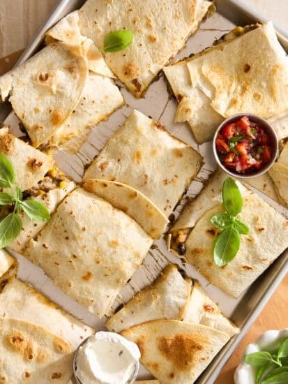 A baking sheet with sliced quesadillas garnished with fresh basil, served with a small bowl of salsa and a dollop of sour cream on the side.