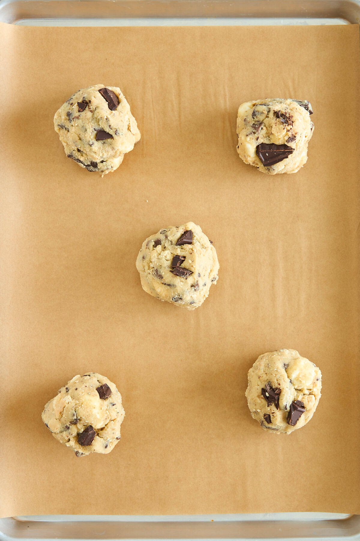 Five balls of chocolate chip cookie dough are evenly spaced on a parchment-lined baking sheet, ready to be baked.