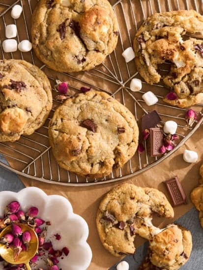 A batch of chocolate chip cookies with chunks of chocolate and marshmallows, some whole and some broken open, are displayed on a wire rack with small marshmallows and rose petals scattered around.