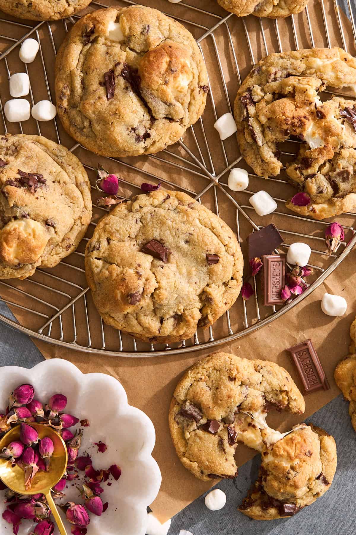 A batch of chocolate chip cookies with chunks of chocolate and marshmallows, some whole and some broken open, are displayed on a wire rack with small marshmallows and rose petals scattered around.