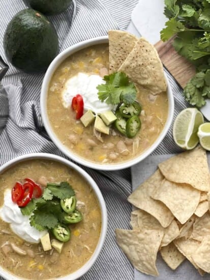 two bowls of white chicken chili with sour cream, avocado, jalapeño, and tortilla chips.