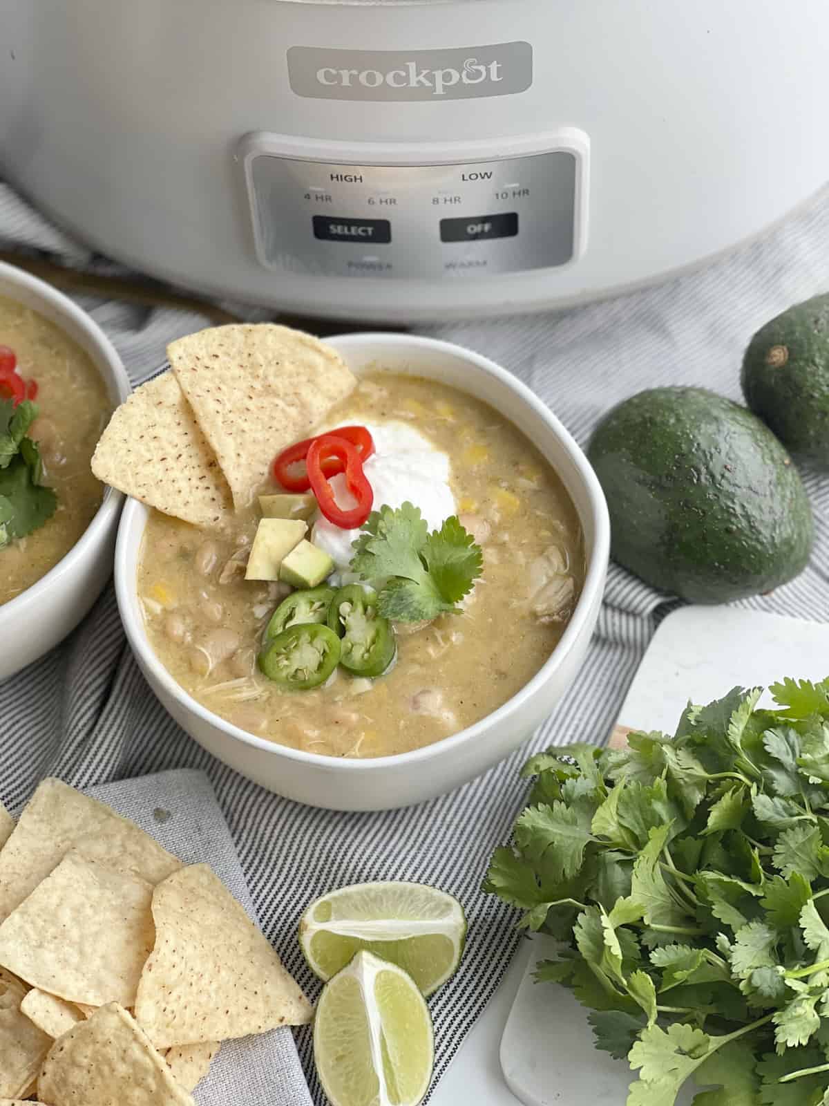 one bowl of white chicken chili with sour cream, avocado, jalapeño, and tortilla chips. pictured with crockpot.
