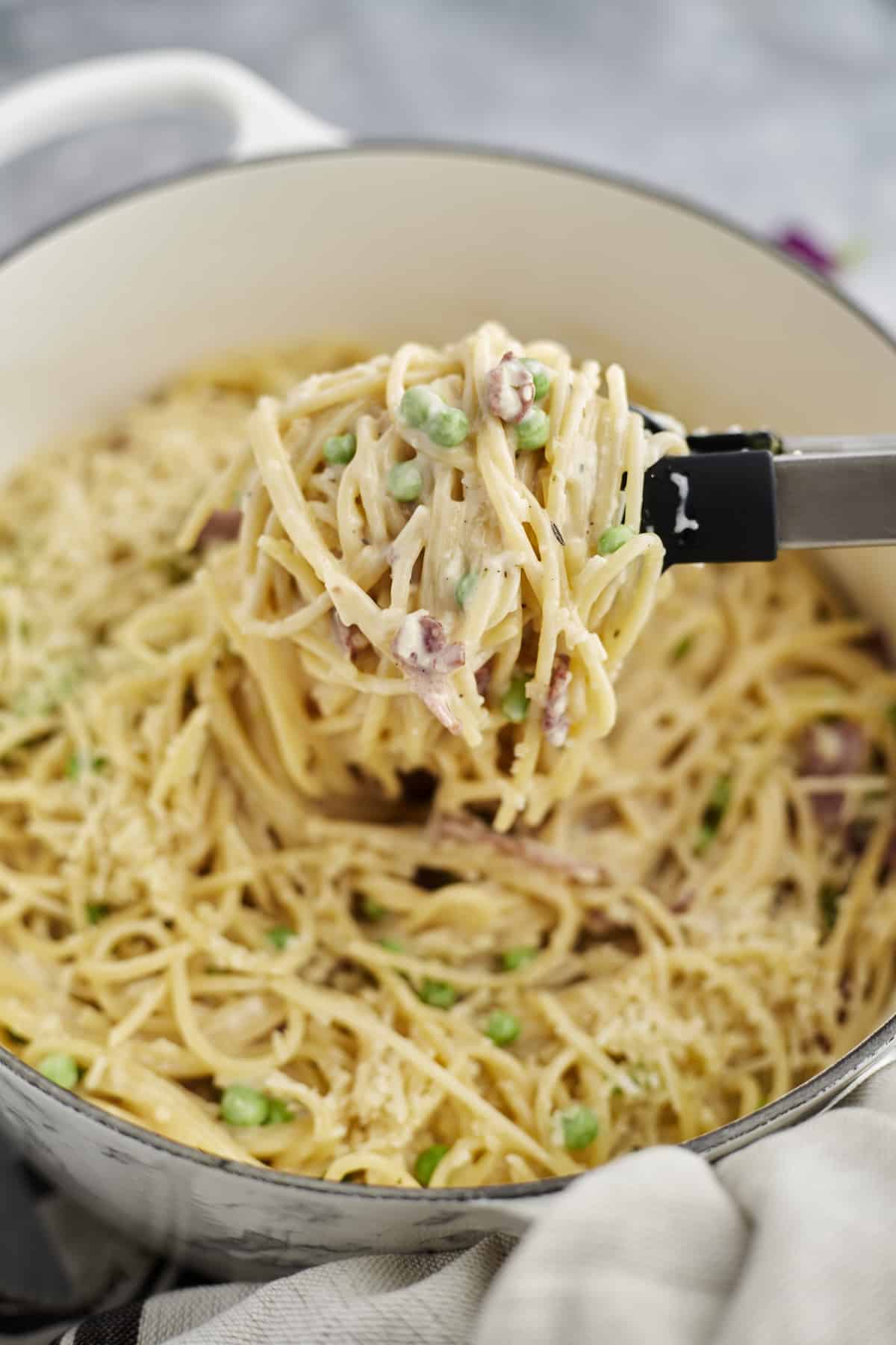 overhead image of spaghetti carbonara in a white pot with a serving being lifted with tongs