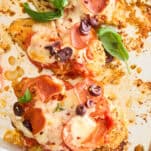 Baked chicken breasts topped with melted cheese, pepperoni slices, black olives, tomato sauce, and fresh basil leaves on a sheet pan.