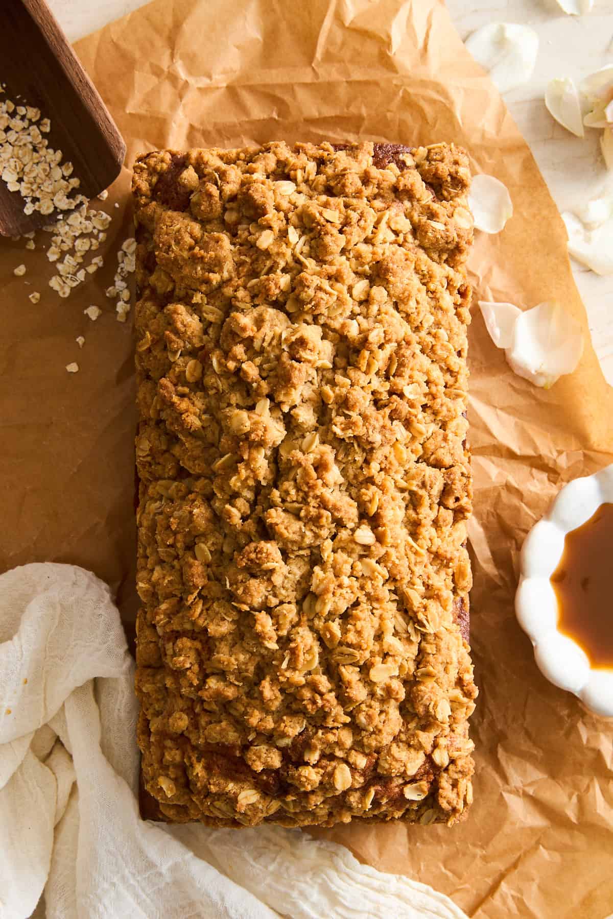 A loaf of bread or cake with a golden oat crumb topping sits on brown parchment paper, surrounded by oats, a white cloth, flower petals, and a small bowl of syrup.