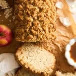 A loaf of apple crumble bread on parchment paper, partially sliced with three pieces cut. An apple, a white cloth, a bowl of caramel sauce, oats, and flower petals are placed around the bread.