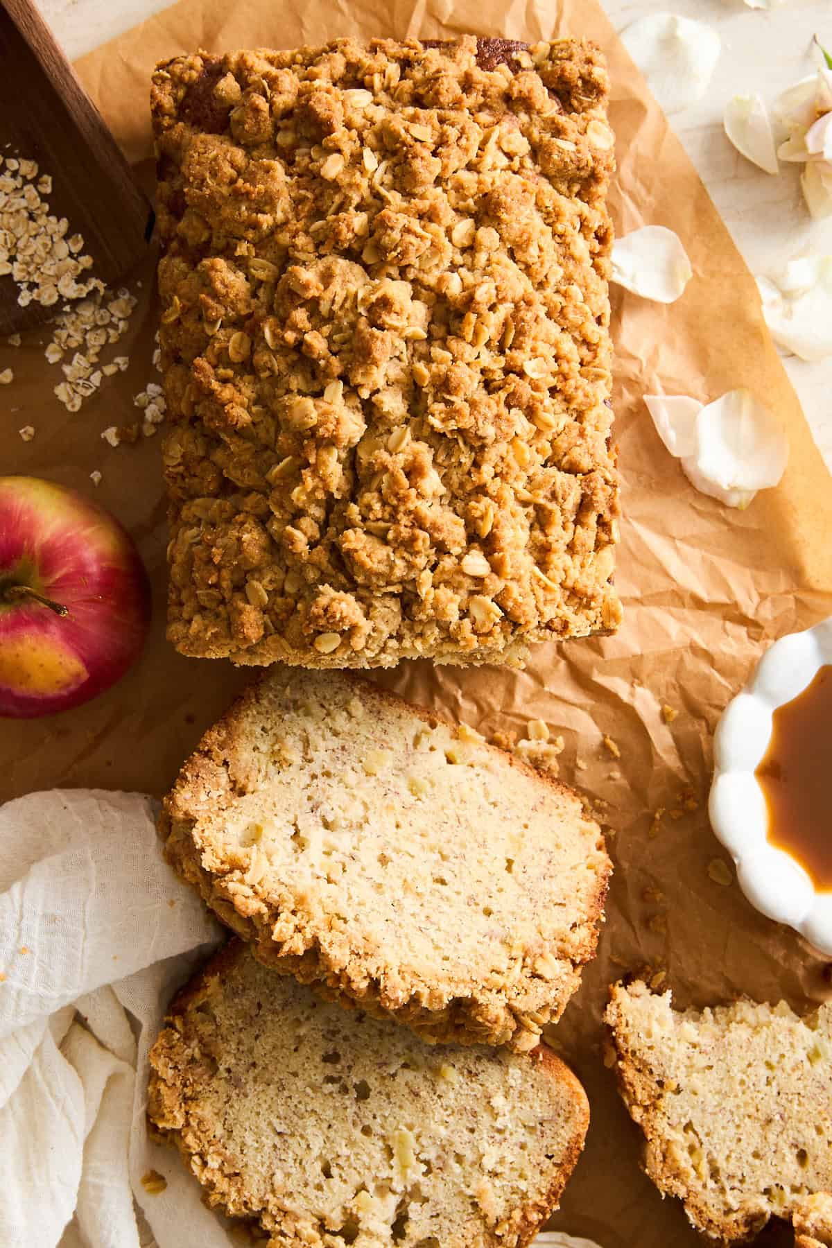 A loaf of apple crumble bread on parchment paper, partially sliced with three pieces cut. An apple, a white cloth, a bowl of caramel sauce, oats, and flower petals are placed around the bread.