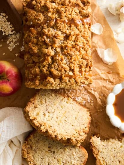 A loaf of crumbly apple bread with a streusel topping sits on brown parchment paper. Three slices are cut, and an apple, oats, a small bowl of caramel, and flower petals are nearby.