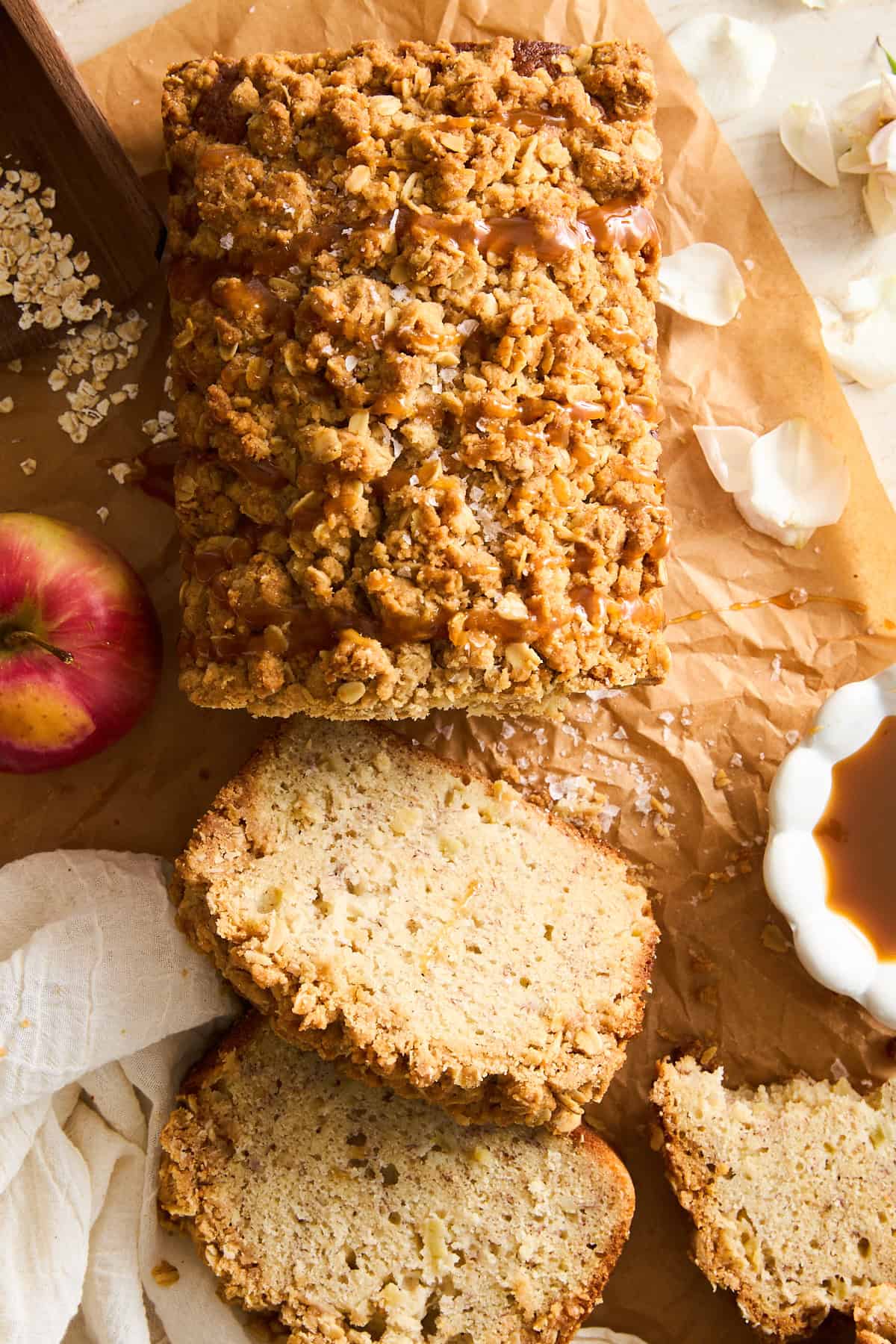 A loaf of crumbly apple bread with a streusel topping sits on brown parchment paper. Three slices are cut, and an apple, oats, a small bowl of caramel, and flower petals are nearby.