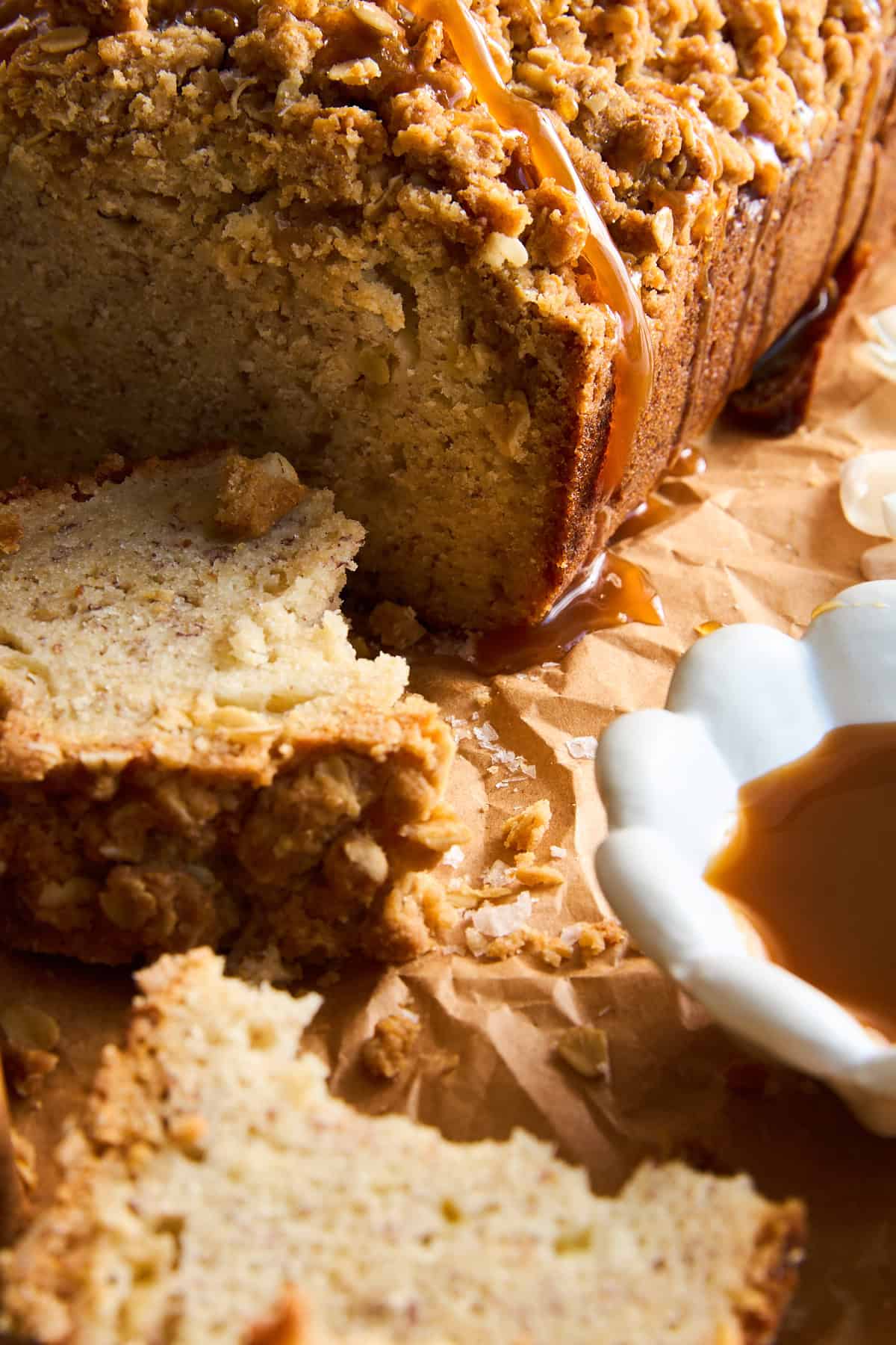 Close-up of sliced crumb cake with a streusel topping and caramel drizzle, resting on brown parchment paper. A white dish with caramel sauce sits nearby. Crumbs are scattered around the slices.